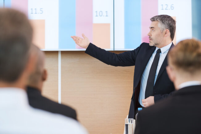 Talking about numbers. Confident mature man in formalwear pointing projection screen with graph on it while making presentation in conference hall with people on the foreground